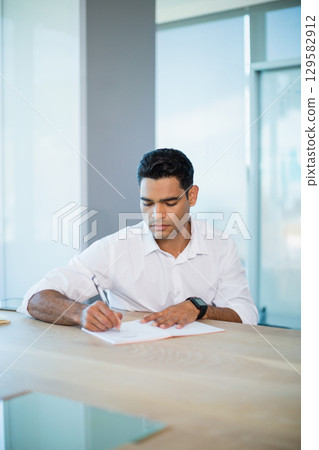 Male office worker writing with pen on paper at office desk wearing white shirt and smartwatch Male office worker writing with pen on paper at office desk wearing white shirt and smartwatch 129582912
