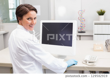 Female researcher wearing lab coat and gloves sitting at lab bench smiling by computer DNA model 129582919