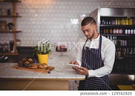 Man wearing striped apron using tablet at cafe counter with pastries, drink display, copy space Man wearing striped apron using tablet at cafe counter with pastries, drink display, copy space 129582920
