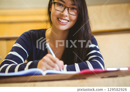 Asian female student smiling and writing in notebook in lecture hall with red tablet on desk 129582926