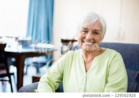 Senior woman sitting in armchair at home by dining table set, smiling at camera, copy space 129582928