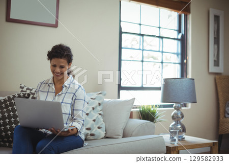 Woman sitting on light fabric sofa in bright living room typing on silver laptop, copy space 129582933