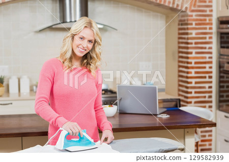 Woman ironing shirt with light blue iron on home kitchen island with laptop and fruit bowl Woman ironing shirt with light blue iron on home kitchen island with laptop and fruit bowl 129582939