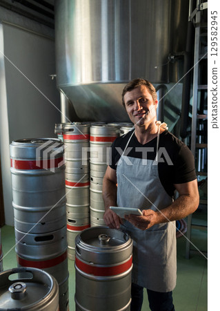 Senior man wearing apron and holding tablet by stainless fermenter tank among kegs in brewery Senior man wearing apron and holding tablet by stainless fermenter tank among kegs in brewery 129582945
