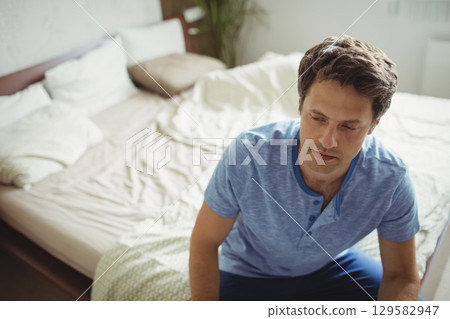 Mid-adult man sitting on bed edge in bedroom wearing blue henley and dark pants, copy space Mid-adult man sitting on bed edge in bedroom wearing blue henley and dark pants, copy space 129582947