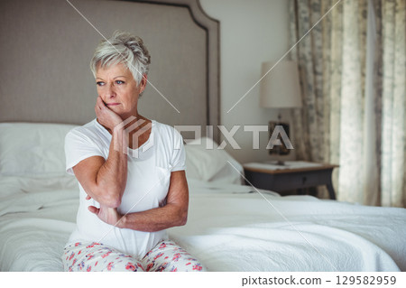 Senior woman resting cheek on hand and crossing arms on bed with white bedding beside lamp Senior woman resting cheek on hand and crossing arms on bed with white bedding beside lamp 129582959