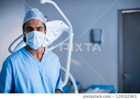 Asian male surgeon standing under overhead lamp in surgery room by table wearing scrubs, copy space 129582963