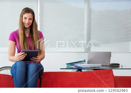 Woman sitting on red sofa holding tablet in home study with laptop, phone, copy space Woman sitting on red sofa holding tablet in home study with laptop, phone, copy space 129582965