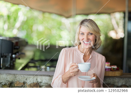 Middle-aged woman wearing pink top smiling holding cup, saucer at cafe with pastries, copy space 129582966