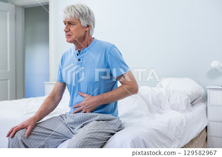 Senior man clutching abdomen while sitting on bed edge in tidy bedroom with nightstand lamp 129582967