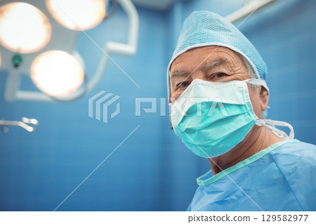Senior male surgeon standing in operating room wearing cap mask under light near clamp, copy space Senior male surgeon standing in operating room wearing cap mask under light near clamp, copy space 129582977