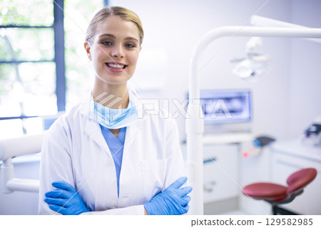 Female dentist standing with arms crossed in dental clinic showing dental chair and instrument tray 129582985