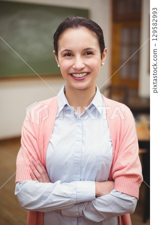Smiling female teacher wearing blue shirt pink cardigan classroom chalkboard, desks, chairs, door Smiling female teacher wearing blue shirt pink cardigan classroom chalkboard, desks, chairs, door 129582993