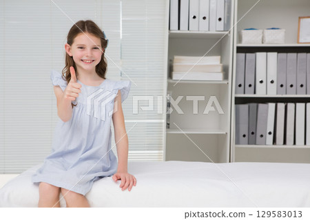 Girl giving thumbs-up sitting on exam table in clinic with blinds and binder-box shelf, copy space 129583013