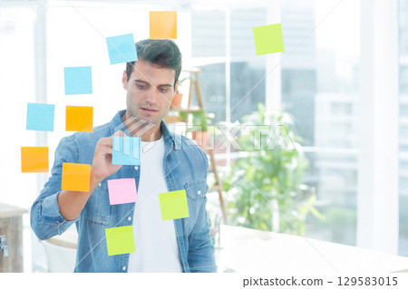 Man wearing denim shirt placing blue sticky note on glass partition in modern office Man wearing denim shirt placing blue sticky note on glass partition in modern office 129583015