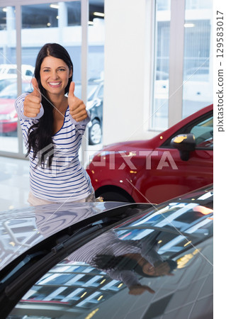 Woman standing next to shiny car bonnet and red hatchback at dealership showroom, giving thumbs-up Woman standing next to shiny car bonnet and red hatchback at dealership showroom, giving thumbs-up 129583017