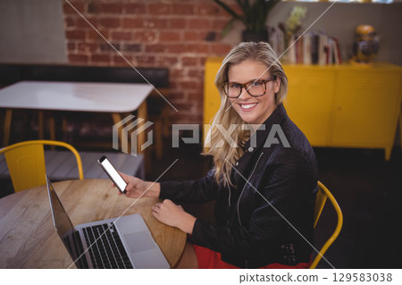 Woman wearing glasses and leather jacket sitting at table in cafe holding smartphone and laptop 129583038