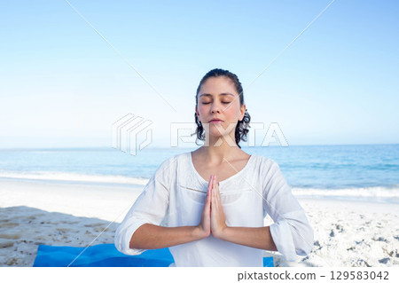 Woman sitting cross-legged on blue yoga mat at shoreline meditating beside calm ocean horizon 129583042