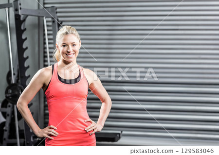 Woman standing with hands on hips at gym beside weighted squat rack and roller shutter door 129583046
