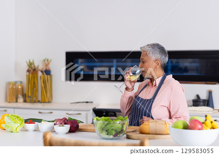Senior Asian woman wearing apron at kitchen island sipping wine from glass and chopping vegetables 129583055