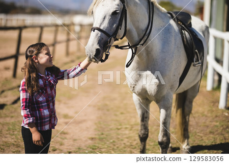 Girl standing beside light gray horse in ranch paddock petting muzzle near wooden rail fence Girl standing beside light gray horse in ranch paddock petting muzzle near wooden rail fence 129583056