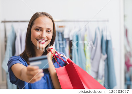 Female shopper smiling, holding credit card and red bags by metal rack in clothing boutique 129583057