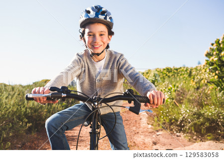 Boy wearing polka-dot helmet riding bicycle along sunlit dirt trail between shrubs under clear sky 129583058