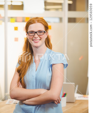 Red-haired woman standing crossing arms in office by glass partition with sticky notes and laptop 129583059