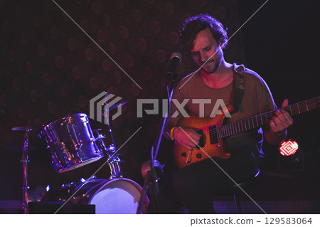 Male musician sitting on stool on club stage playing guitar singing into mic, copy space 129583064