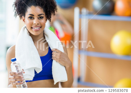 African American woman holding water bottle near exercise ball rack in gym wearing blue sportswear African American woman holding water bottle near exercise ball rack in gym wearing blue sportswear 129583078