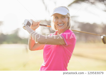 Senior woman swinging golf club on fairway wearing pink polo, white visor and glove, copy space 129583079