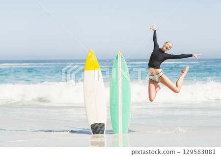 Female surfer leaping in shallow water at sandy beach near two surfboards, copy space Female surfer leaping in shallow water at sandy beach near two surfboards, copy space 129583081