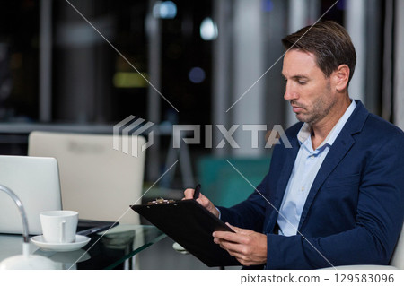 Businessman sitting at glass-top desk in office taking notes on clipboard near laptop coffee cup Businessman sitting at glass-top desk in office taking notes on clipboard near laptop coffee cup 129583096