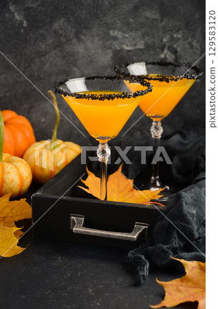 Fall seasonal cocktail featuring vibrant pumpkin martini in elegant glasses with black rim, surrounded by autumn leaves and decorative pumpkins 129583120