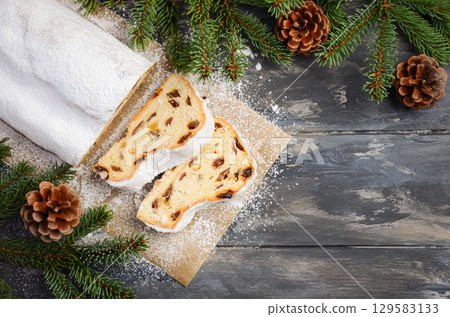 Christmas Stollen loaf with powdered sugar, sliced to reveal rich fruit and nut filling, surrounded by festive pine branches and pine cones 129583133