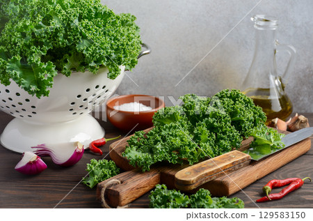 Fresh green kale leaves arranged on a wooden cutting board with olive oil, salt, and red chili peppers in a rustic kitchen setting Fresh green kale leaves arranged on a wooden cutting board with olive oil, salt, and red chili peppers in a rustic kitchen setting 129583150