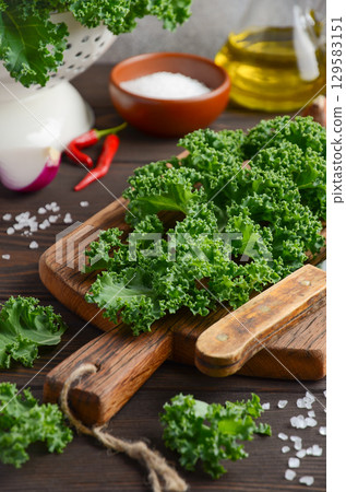 Fresh green kale leaves arranged on wooden cutting board with olive oil, salt, and red chili peppers in a rustic kitchen setting Fresh green kale leaves arranged on wooden cutting board with olive oil, salt, and red chili peppers in a rustic kitchen setting 129583151