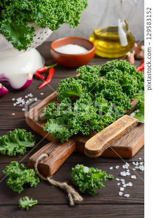 Fresh green kale leaves arranged on a wooden cutting board with salt, olive oil, and red chili peppers in a rustic kitchen setting Fresh green kale leaves arranged on a wooden cutting board with salt, olive oil, and red chili peppers in a rustic kitchen setting 129583152