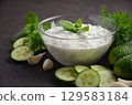 Creamy Greek dip sauce tzatziki in a glass bowl surrounded by fresh cucumbers, garlic cloves, and herbs on a dark textured surface 129583184