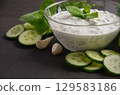 Creamy Greek dip sauce tzatziki in a glass bowl garnished with fresh mint leaves, surrounded by sliced cucumbers and garlic cloves, perfect for appetizers 129583186