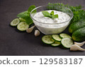Creamy Greek dip sauce tzatziki in a glass bowl surrounded by fresh cucumbers, garlic, and herbs, perfect for enhancing Mediterranean dishes 129583187