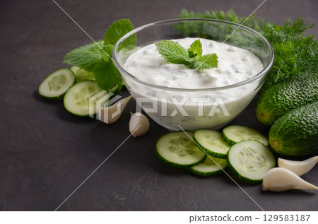 Creamy Greek dip sauce tzatziki in a glass bowl surrounded by fresh cucumbers, garlic, and herbs, perfect for enhancing Mediterranean dishes 129583187