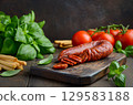 Sliced spicy Chorizo sausage on wooden board surrounded by fresh tomatoes, basil leaves, and breadsticks, showcasing vibrant culinary ingredients and textures 129583188