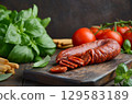 Sliced cured Chorizo sausage on wooden cutting board surrounded by fresh basil leaves and ripe tomatoes, showcasing vibrant colors and culinary ingredients 129583189