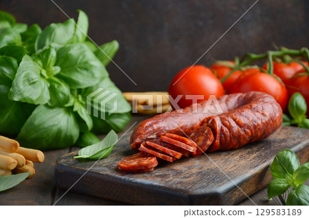 Sliced cured Chorizo sausage on wooden cutting board surrounded by fresh basil leaves and ripe tomatoes, showcasing vibrant colors and culinary ingredients Sliced cured Chorizo sausage on wooden cutting board surrounded by fresh basil leaves and ripe tomatoes, showcasing vibrant colors and culinary ingredients 129583189
