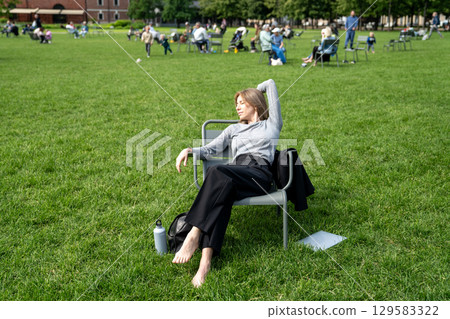 Work-weary woman take break relaxed reclines on chair in open public space on lawn hands over eyes. Work-weary woman take break relaxed reclines on chair in open public space on lawn hands over eyes. 129583322