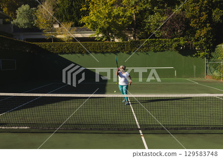 Senior African American woman in sportswear swinging racket preparing to hit tennis ball on court 129583748