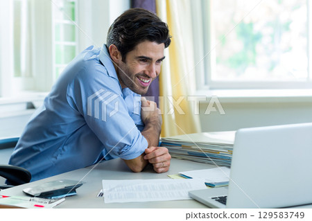 Mid adult man smiling at silver laptop while tapping smartphone then sorting file folders at office 129583749