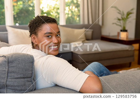 African American adult woman reclining on grey sofa by tall windows at home with folded book 129583756