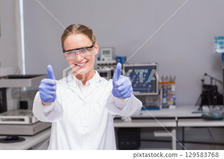 Woman scientist standing in lab wearing lab coat goggles gloves and giving thumbs up 129583768